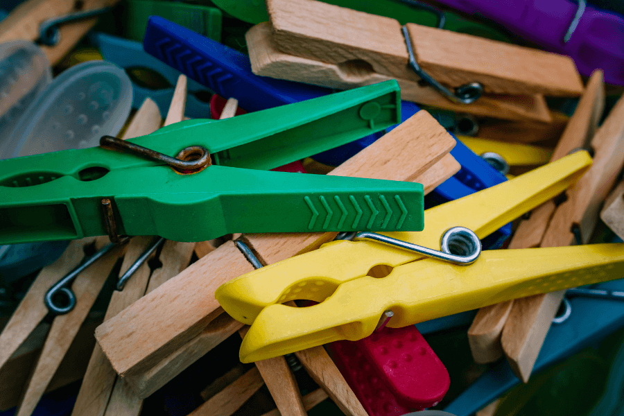 The image is a close-up of a colorful assortment of clothespins, including green, yellow, wooden, and purple varieties, scattered in a pile. The vibrant colors and different textures of the clothespins create a visually engaging scene, emphasizing everyday objects with a focus on utility and variety.