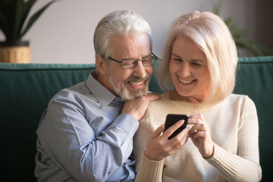 Mature attractive couple sitting on couch using smartphon