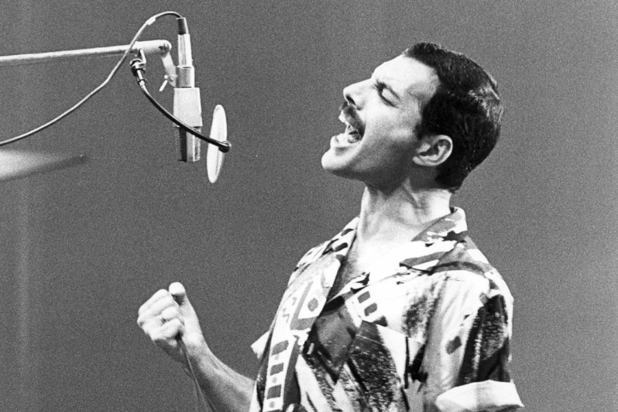 A black and white photograph of a male singer passionately performing in front of a studio microphone. He wears a patterned, short-sleeved button-up shirt, tilts his head back, and sings with emotion, his fist clenched. The lighting highlights his expressive face and strong posture.