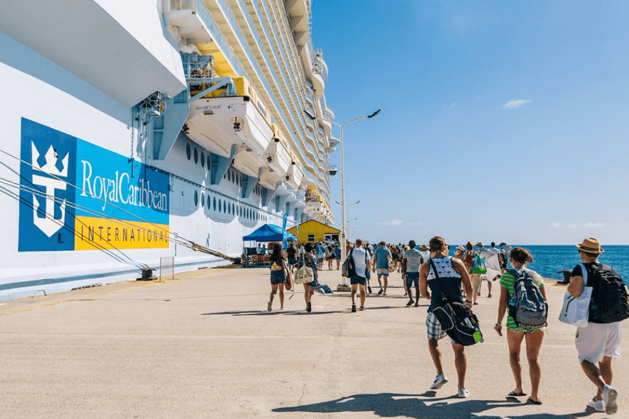 A large Royal Caribbean cruise ship docked at a sunny port with a crowd of passengers walking along the pier. The ship's blue and yellow logo is visible on the side, and people dressed in casual summer clothing, some carrying backpacks and beach bags, head towards the shore. The ocean stretches out in the background under a bright blue sky.