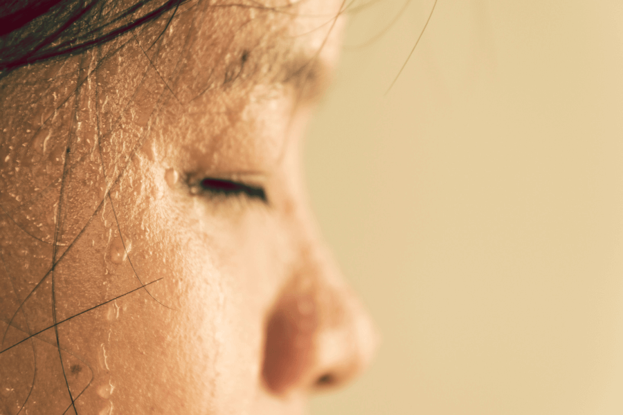 Close-up of a person's face with closed eyes, covered in sweat droplets. The warm lighting and soft focus highlight the texture of the skin and the moisture on the surface.