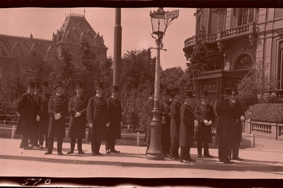 A sepia-toned historical photograph showing a group of men dressed in long black coats, white gloves, and tall top hats, standing in a formal arrangement on a sidewalk. They appear to be engaged in a ceremonial or official gathering near a lamppost with a street sign that reads "TURFMARKT." The backdrop features a grand European-style building with ornate balconies and a steep-roofed structure, suggesting a late 19th or early 20th-century setting.