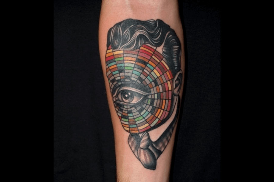A vibrant tattoo of a surrealist design featuring a man's profile with slicked-back hair and a pipe. The face is replaced with a colorful radial pattern resembling a color wheel, with a single, detailed eye in the center.