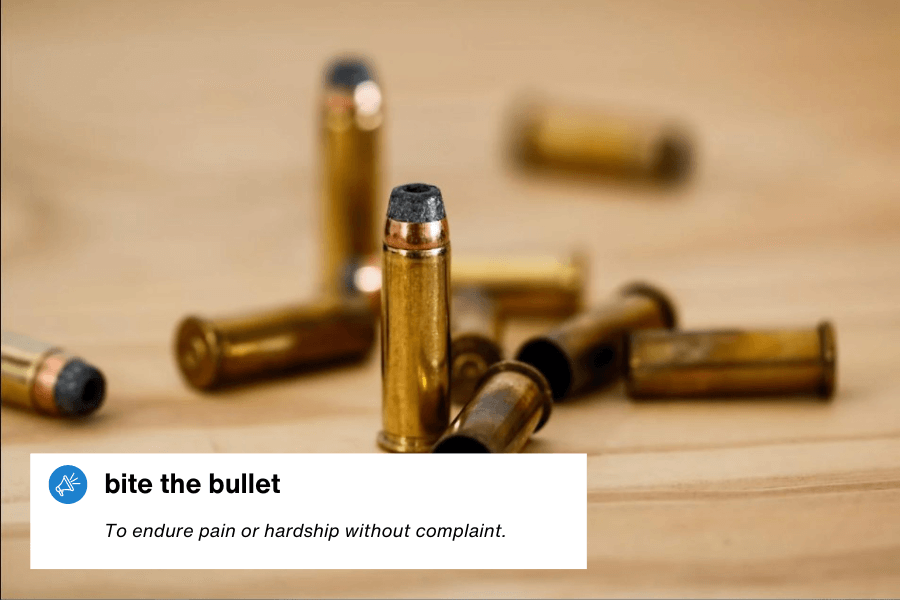 A close-up of several bullets scattered on a wooden surface, with one standing upright in the center. A text overlay in the bottom left corner reads: "Bite the bullet – To endure pain or hardship without complaint."