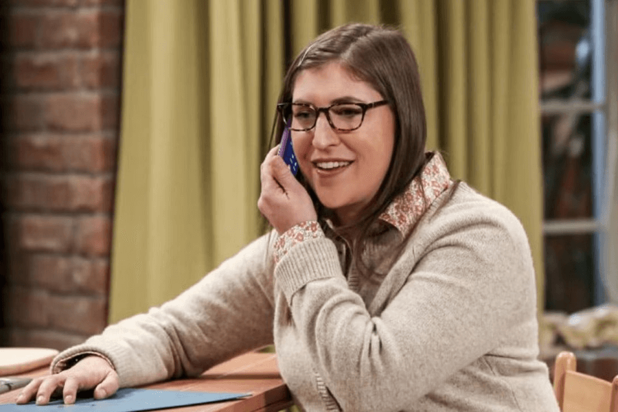 A woman with long brown hair, glasses, and a warm smile is sitting at a table while talking on a cellphone. She wears a beige cardigan over a patterned collared shirt and appears engaged in conversation. The background features a brick wall and green curtains, suggesting a cozy indoor setting.