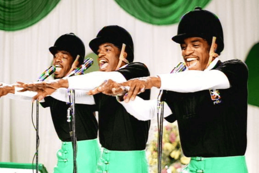 Three performers dressed in matching black shirts, green pants, and black equestrian helmets sing and dance in unison on a stage with green and white drapery. They each have a drumstick tucked behind their ear and are reaching forward with animated expressions. The image appears to be a still from a music video or performance.