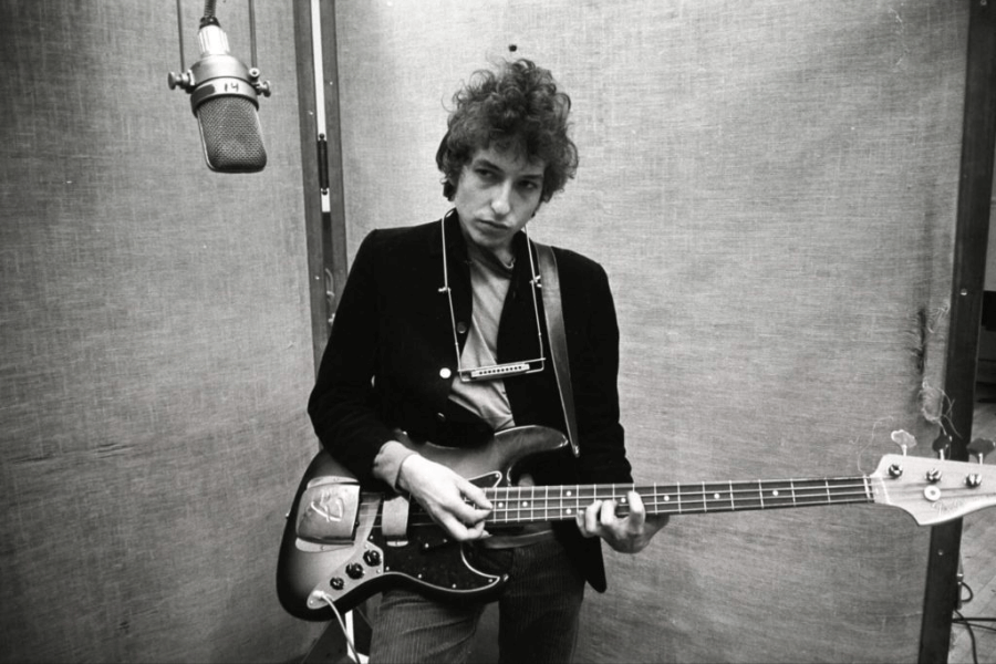  A black and white photograph of a male musician standing in a recording studio, holding a bass guitar with a harmonica around his neck. He wears a dark jacket over a simple shirt, his curly hair slightly unkempt, and his expression appears introspective. A vintage microphone hangs in front of him, enhancing the raw and intimate atmosphere of the moment.