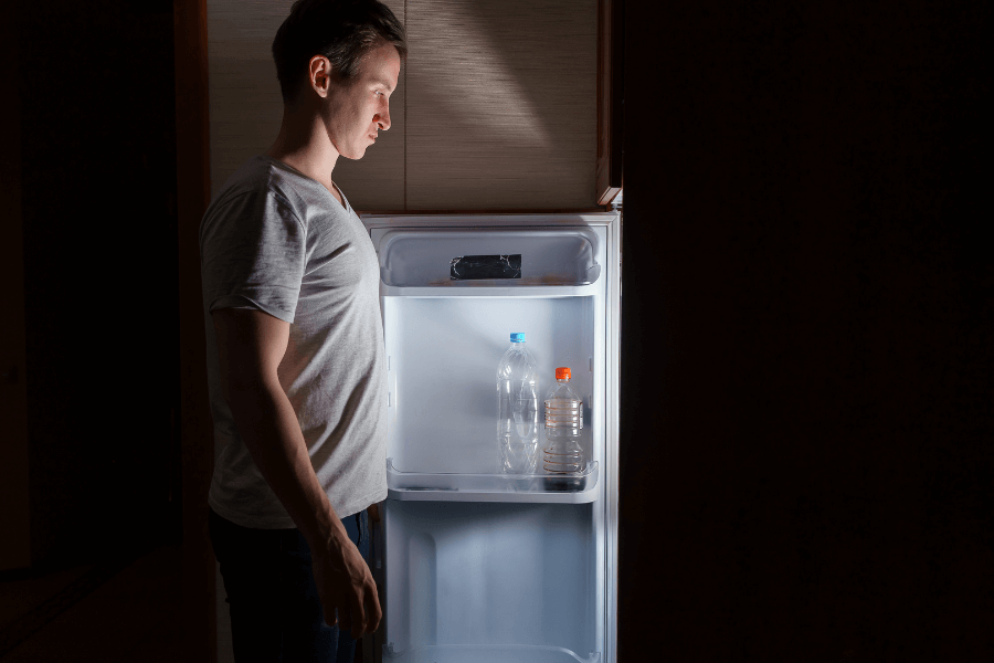 A man in a white t-shirt stands in the dark, staring into an almost empty refrigerator with only a few bottles inside. The fridge’s light illuminates his face, highlighting his disappointed or thoughtful expression.