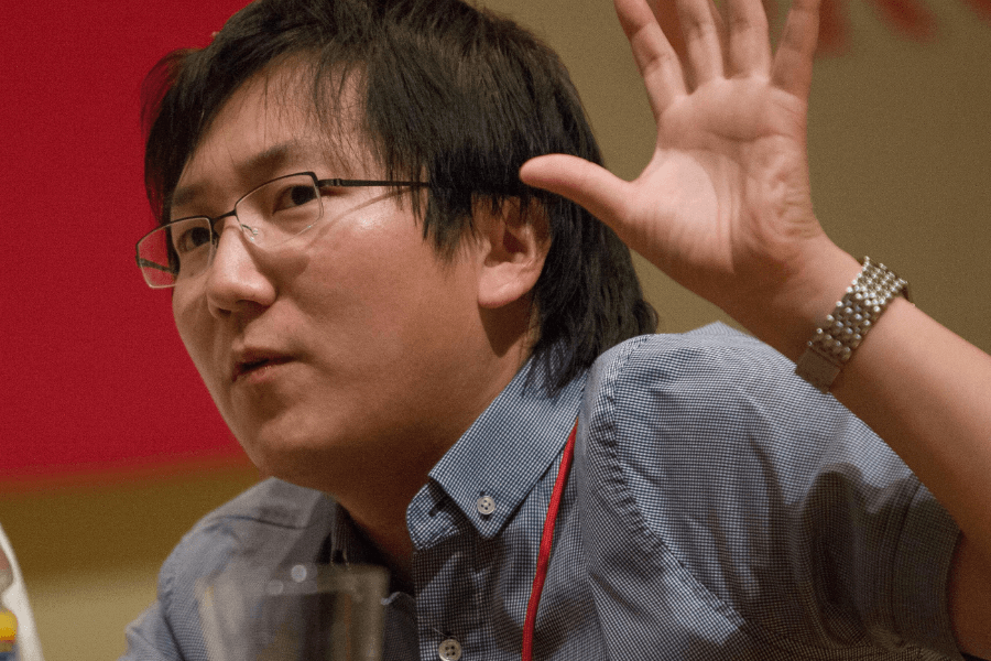 A man with medium-length black hair and glasses is mid-conversation, raising his hand expressively. He wears a blue button-up shirt with a red lanyard around his neck and a silver watch on his wrist. The background is blurred, featuring warm indoor lighting and red and beige tones.