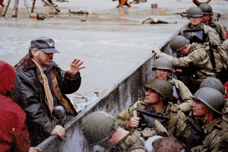 A film director in a navy cap and dark jacket gestures while giving instructions to a group of actors dressed as World War II soldiers. The soldiers, wearing helmets and holding rifles, stand inside a landing craft, recreating a beach invasion scene. The wet, overcast setting and props suggest a historically inspired war film production.