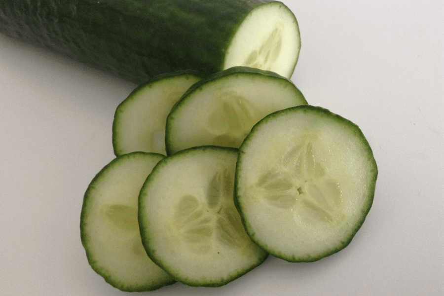 A fresh cucumber partially sliced on a white surface, with several round pieces arranged in the foreground. The green skin contrasts with the pale, juicy interior filled with seeds.