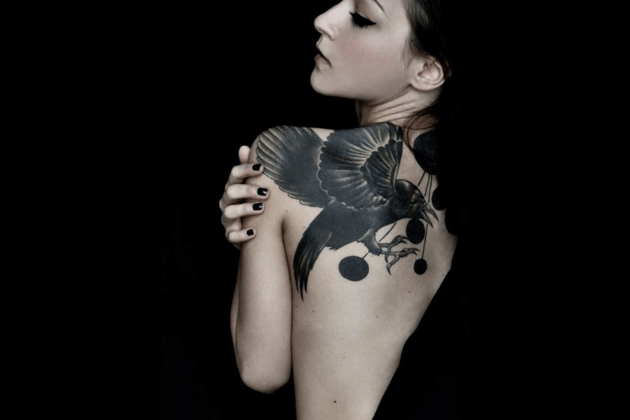 A striking black tattoo of a raven on a woman's upper back. The raven is detailed, mid-flight, clutching small circular objects in its talons, with its wings spread wide.