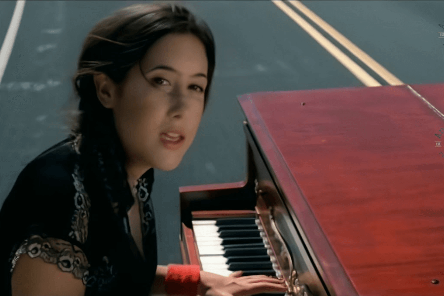 A young woman with dark hair plays a wooden grand piano while looking towards the camera, appearing to sing. The piano is set on an open road with yellow dividing lines, giving the scene a surreal and cinematic feel. The motion blur in the background suggests movement, as if the piano is gliding down the street.