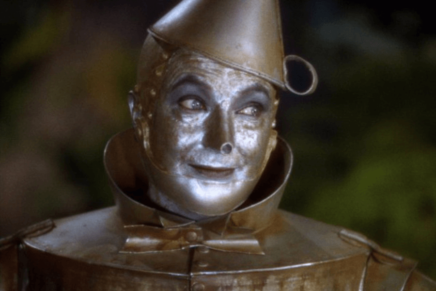 A metallic-looking character with a silver-painted face and a conical hat smiles subtly while looking off to the side. He wears a structured, tin-like suit with a bowtie, giving him a mechanical yet expressive appearance. The soft lighting and blurred background suggest a whimsical or fantastical setting.