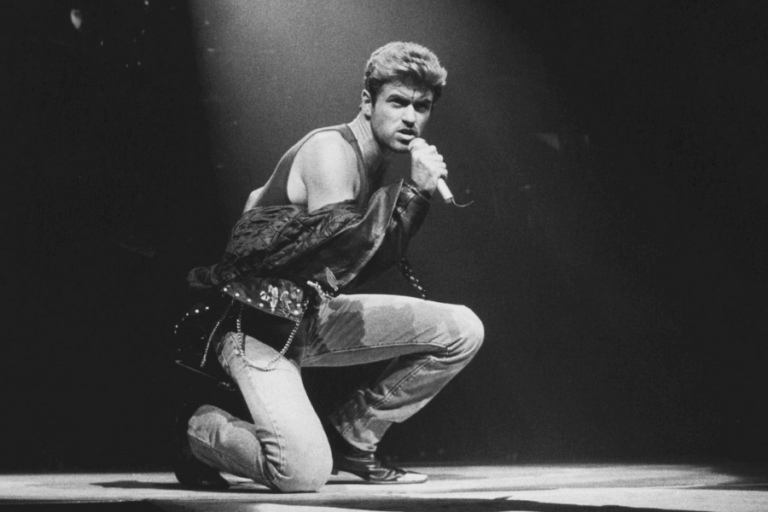 A black and white photograph of a male singer mid-performance, crouching on stage while gripping a microphone. He wears a sleeveless shirt, distressed jeans, and a partially unzipped leather jacket draped over his shoulders, exuding a rebellious and charismatic stage presence. A spotlight highlights his intense gaze as he engages with the audience.