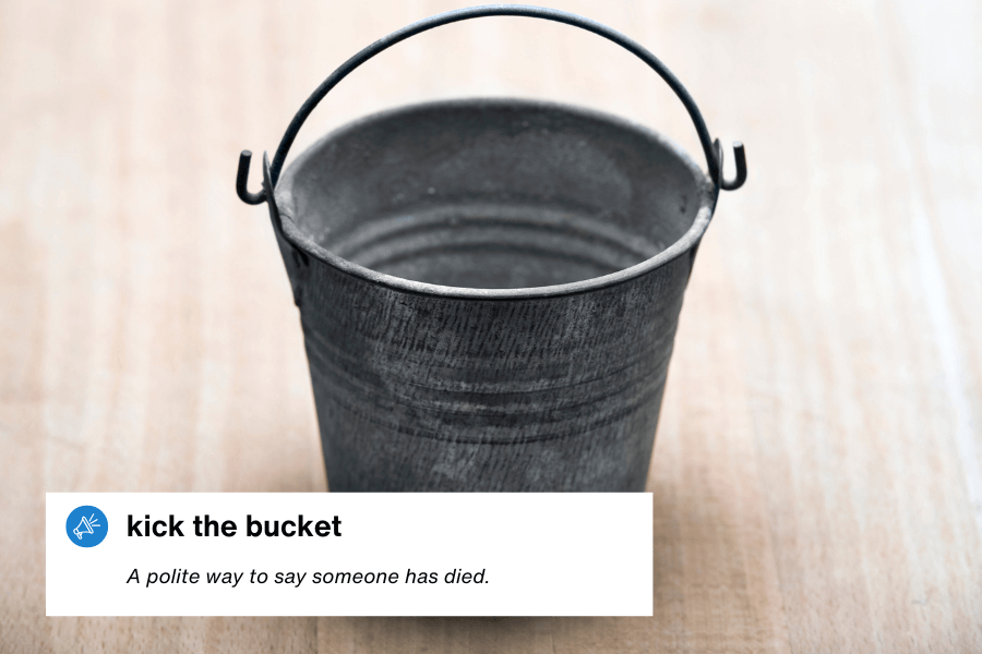 A close-up photograph of a small, empty metal bucket with a curved handle, sitting on a wooden surface. A text overlay in the bottom left corner reads: "Kick the bucket – A polite way to say someone has died."