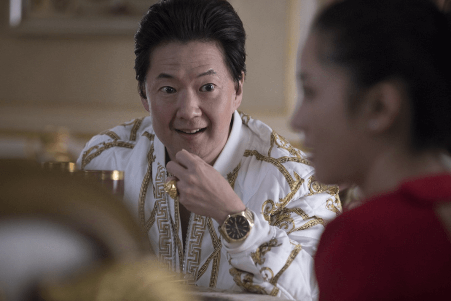 A man wearing an ornate white and gold jacket sits at a luxurious dining table, speaking animatedly with a woman in a red outfit who is slightly out of focus in the foreground. He has a confident and amused expression, accessorized with a large gold ring and a stylish watch. The opulent setting and his expressive demeanor suggest a scene of wealth and humor.