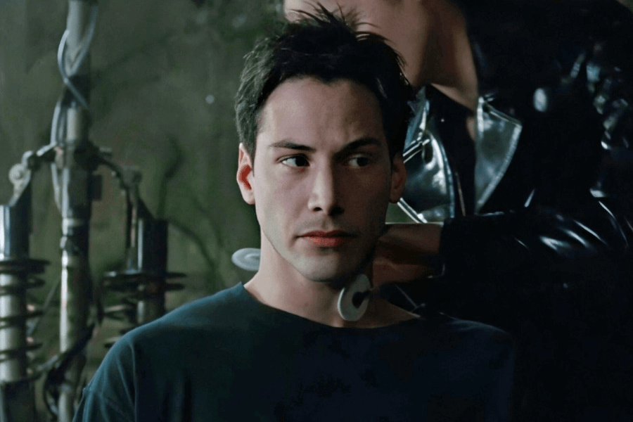 A young man with dark hair and a serious expression sits in a dimly lit, industrial-style room. A person in a black leather jacket is behind him, attaching a circular device to the back of his neck. The scene has a futuristic, cyberpunk atmosphere, with exposed wires and mechanical components in the background.
