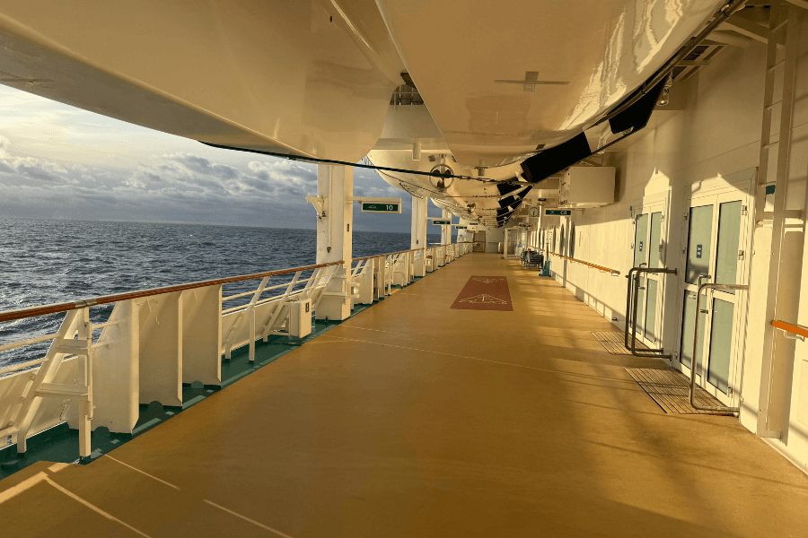 A serene cruise ship promenade deck bathed in golden sunlight, with the vast ocean stretching out to the horizon. The deck is lined with white railings, emergency doors, and lifeboats overhead, creating a peaceful and structured walkway. The calm sea and scattered clouds in the background add to the tranquil atmosphere.