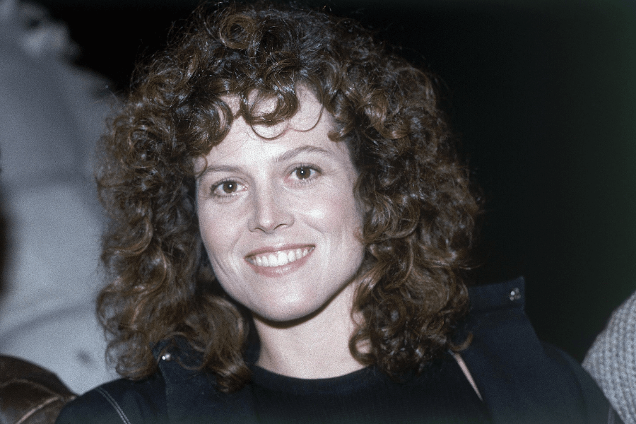 A woman with voluminous curly brown hair smiles warmly at the camera. She is wearing a black top with a dark jacket, and the dimly lit background suggests a nighttime or indoor event. Her natural and radiant expression exudes confidence and charm.