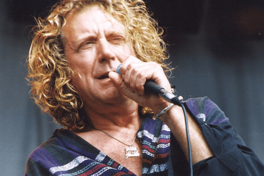  A male rock singer with curly blonde hair passionately performs on stage, gripping a microphone close to his mouth. He wears a loose, patterned shirt with a deep neckline, accessorized with a silver bracelet and a pendant necklace. His intense facial expression and slightly squinting eyes capture the raw energy of the performance.