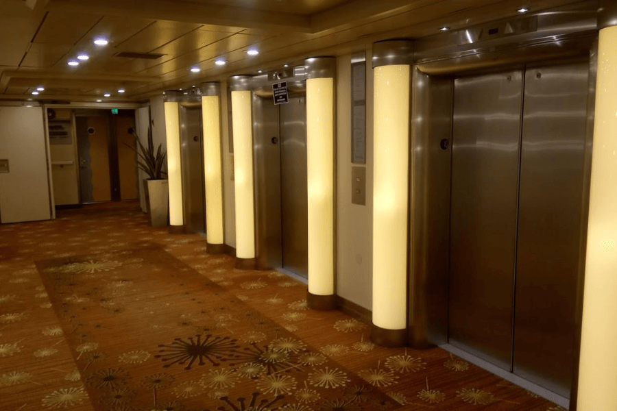 A well-lit cruise ship elevator lobby featuring multiple stainless steel elevator doors framed by tall, glowing light columns. The floor is covered in a patterned carpet with warm tones, complementing the soft ceiling lighting. A potted plant is positioned near the far wall, adding a touch of decor to the modern and clean space.