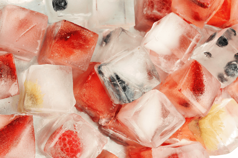 A pile of ice cubes with frozen fruits inside, including strawberries, blueberries, raspberries, and citrus pieces. The vibrant colors of the fruit contrast with the clear and frosty ice.