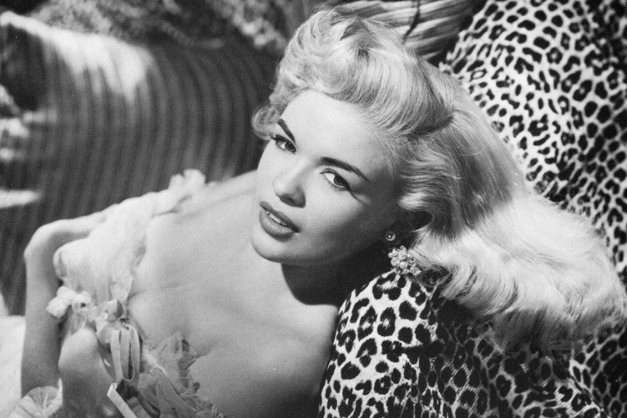 A glamorous woman with voluminous, wavy blonde hair reclines against a leopard-print cushion in this black-and-white portrait. She wears a delicate, ruffled off-the-shoulder gown and sparkling earrings, exuding a classic Hollywood allure. Her sultry expression and elegant styling reflect the golden age of cinema.