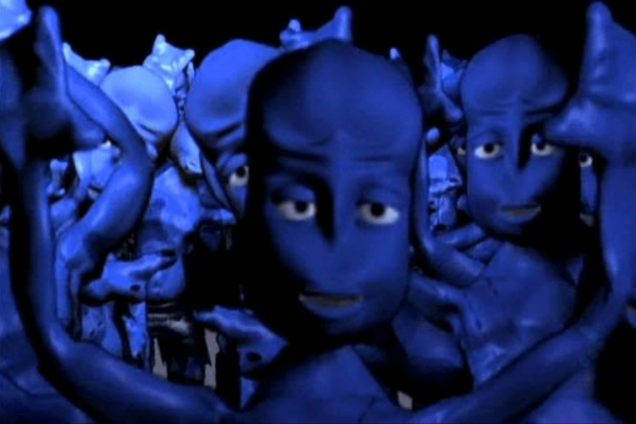 A group of surreal, animated blue humanoid figures with exaggerated facial features and sleepy expressions stand close together in a dark setting. Their fluid, rubbery appearance and synchronized hand gestures suggest a stylized, otherworldly aesthetic. The image appears to be from a CGI-based music video or animation.