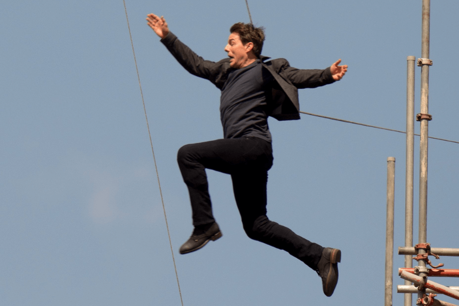 A man in a dark suit leaps through the air with his arms spread wide and an intense expression on his face. He is mid-jump between two structures, with scaffolding visible to his right and a clear blue sky behind him. The scene captures the thrilling moment of an action stunt in progress.