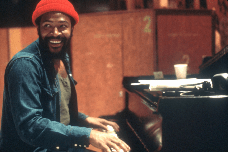 A male musician sits at a grand piano, smiling warmly while playing. He wears a red beanie, a denim jacket over a dark tank top, and has a neatly groomed beard. The setting appears to be a recording studio, with numbered soundproof panels and scattered papers on the piano.