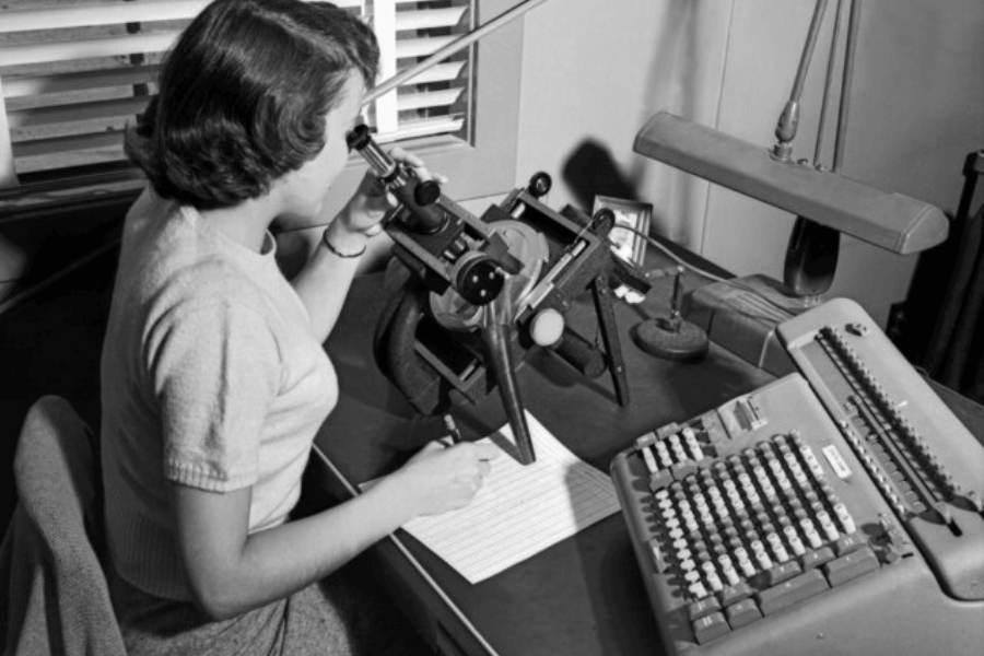 A black-and-white photograph of a woman working at a desk, looking through a microscope-like device while taking notes on a sheet of paper. Beside her is an early mechanical calculator with rows of buttons, suggesting a mid-20th-century office or scientific setting. The scene is illuminated by a desk lamp, and the blinds in the background cast shadows on the wall.
