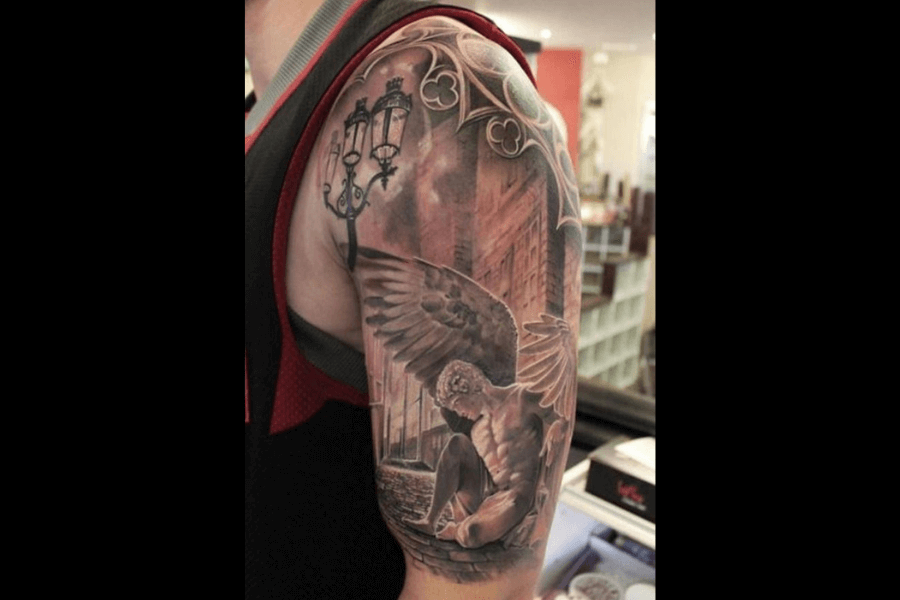 A highly detailed tattoo of a fallen angel with large wings, sitting on cobblestone streets. The design incorporates gothic elements, such as ornate lamp posts, arches, and a dramatic cloudy background.
