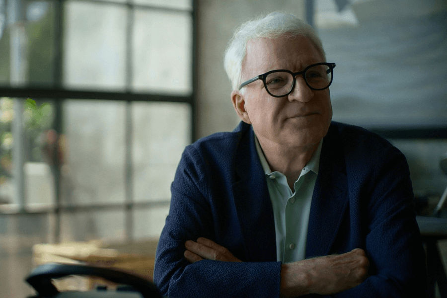 An older man with white hair, black-framed glasses, and a gentle smile sits with his arms crossed in a softly lit modern space. He wears a navy blazer over a light green button-up shirt, and the background features large industrial-style windows with natural light filtering in. His relaxed expression and thoughtful gaze create a warm and introspective atmosphere.