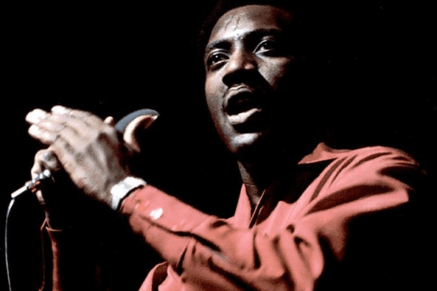 A male soul singer passionately performs on stage, wearing a deep red shirt with sleeves rolled up. He grips the microphone while clapping his hands, his face glistening with sweat and filled with raw emotion. The dark background emphasizes his intense presence under the stage lighting.