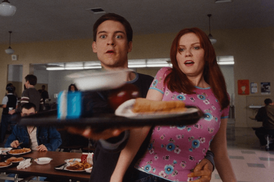 A young man with a surprised expression reaches out with one hand to catch a cafeteria tray filled with food, including a carton of milk, an apple, and a plate. A red-haired young woman in a pink floral top stands beside him, looking equally shocked as she is slightly held by him. The background shows a high school cafeteria filled with students, adding to the everyday setting turned extraordinary.