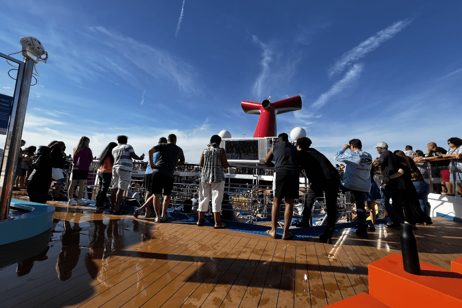  A group of cruise passengers gathers on the open deck of a Carnival cruise ship, enjoying the ocean views and entertainment. The ship's iconic red funnel stands against a bright blue sky with wispy clouds. The wooden deck is partially wet, reflecting sunlight, while guests in casual attire lean against the railing and engage with each other.