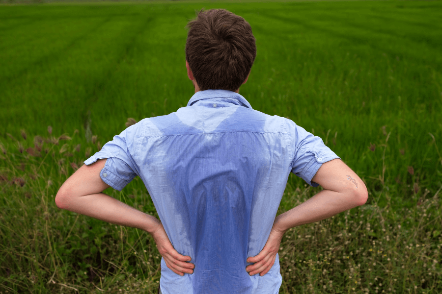 A man wearing a light blue shirt stands with his hands on his hips, facing a vast green field. The back of his shirt is visibly soaked with sweat, suggesting a hot or physically demanding environment.