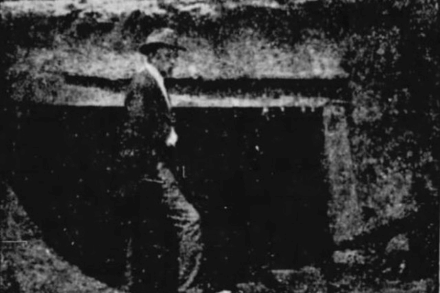 A grainy black-and-white photograph depicting a man in a suit and hat standing in front of a dark, rectangular entrance carved into a rocky hillside. The poor image quality and high contrast obscure finer details, but the setting suggests an archaeological site or an old tomb entrance. The man's posture and placement imply he is examining or presenting the discovery.