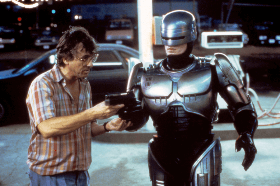 A man in a casual plaid shirt and glasses holds a futuristic handgun while demonstrating something to a robotic police officer in metallic armor. The armored figure, resembling a cyborg, stands rigidly, listening intently in a dimly lit urban setting with a gas station and parked cars in the background. The scene exudes a sci-fi, action-movie atmosphere.