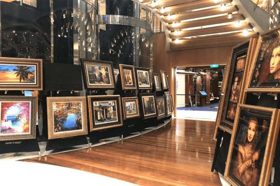 A cruise ship art gallery featuring an elegant display of framed paintings on black easels along a polished wooden floor. The artwork includes colorful landscapes, cityscapes, and portraits, arranged under soft, ambient lighting. In the background, a spiral staircase with glass railings and a warmly lit hallway add to the luxurious atmosphere.