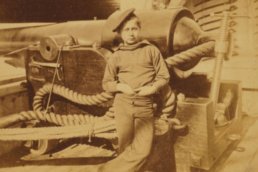 A sepia-toned historical photograph of a young sailor boy leaning casually against a large naval cannon aboard a ship. He wears a loose-fitting sweater, trousers, and a beret, with his hands tucked into his waistband. Thick ropes are coiled around the cannon, and the deck is lined with wooden planks, suggesting a late 19th or early 20th-century maritime setting.
