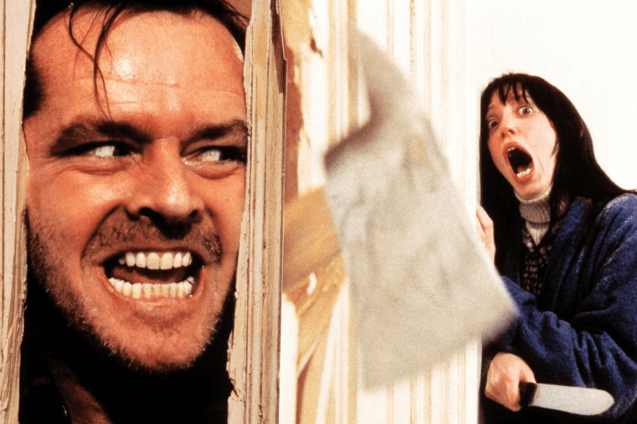 A man with a wild expression and gritted teeth peers through a broken wooden door, his face full of manic energy. On the other side, a terrified woman in a blue robe screams, holding a knife defensively as an axe swings through the air. The scene is filled with tension and horror, capturing an iconic cinematic moment.