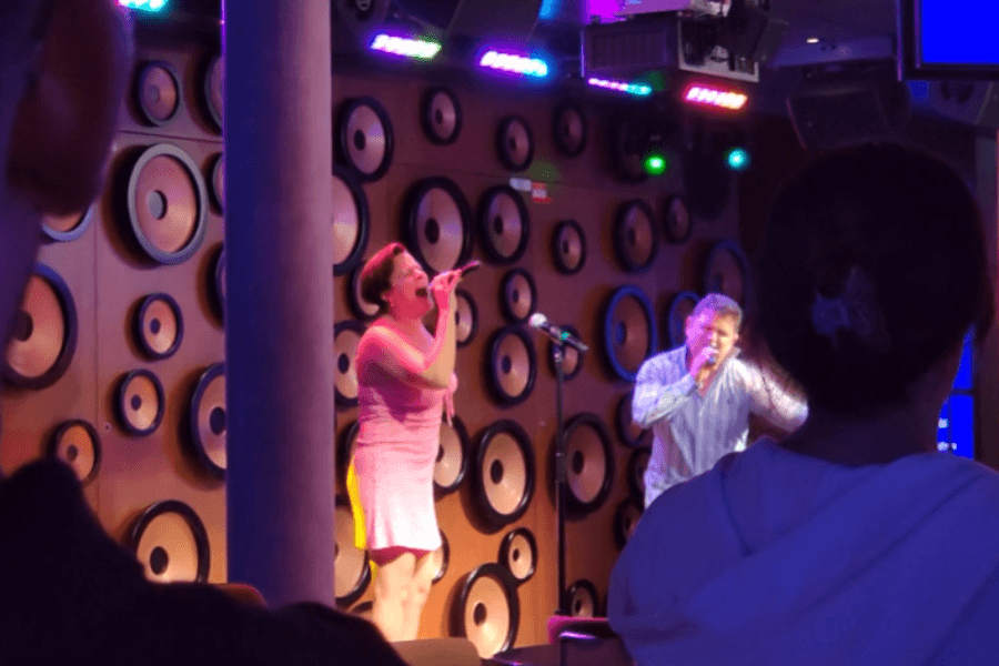 Two cruise passengers passionately perform karaoke on a dimly lit stage with a backdrop featuring a wall of circular speaker-like decorations. The woman in a pink dress sings into the microphone with an expressive gesture, while the man in a striped shirt enthusiastically joins in. Colorful stage lights illuminate the scene, as an engaged audience watches from their seats.