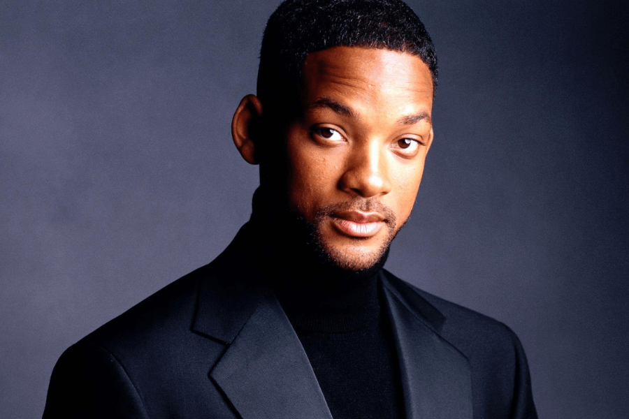 A man with short black hair and a well-groomed beard gazes directly at the camera with a confident yet approachable expression. He wears a sleek black suit over a black turtleneck, set against a softly lit dark gray background. The lighting highlights his facial features, giving the portrait a polished and sophisticated look.