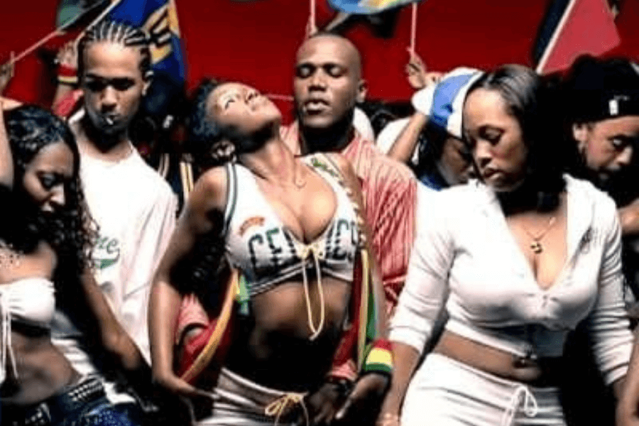 A vibrant party scene featuring a group of people dancing energetically in a crowded space, with flags waving in the background. The central figures include a woman wearing a cropped top with "Cenac" written on it, tilting her head back as she dances, and a man in a red jacket behind her. The lively atmosphere, bold outfits, and Caribbean-inspired elements suggest a dancehall or reggae-themed music video.