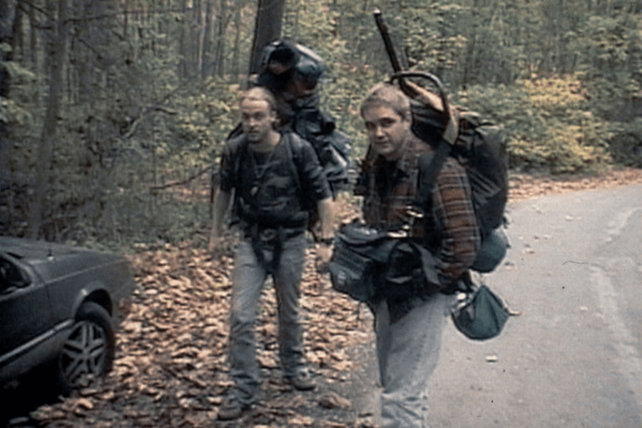 Two hikers with large backpacks and camping gear stand on a leaf-covered forest trail, looking slightly uneasy. One wears a flannel shirt while the other wears a dark t-shirt, and a parked car is visible on the left, partially in the frame. The grainy, handheld-camera aesthetic adds a sense of realism and foreboding.