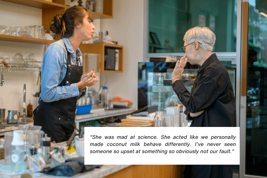 This image shows a barista wearing an apron engaged in a discussion with a customer at a counter in a café. The background includes a shelf with glassware and a refrigerated display case. A quote in the image reads: "She was mad at science. She acted like we personally made coconut milk behave differently. I’ve never seen someone so upset at something so obviously not our fault."