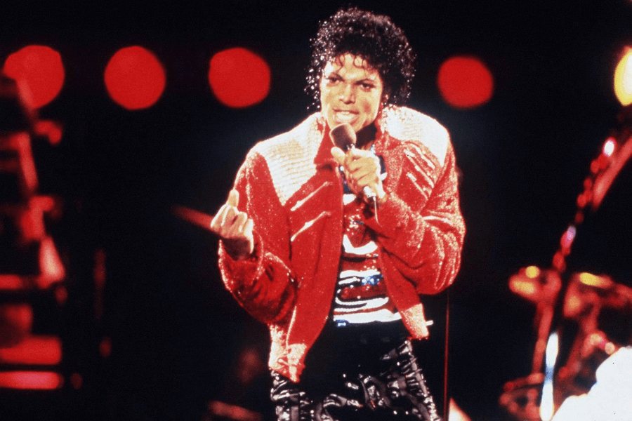 A dynamic concert photograph of a male performer dressed in a sparkling red and white jacket, black pants, and a sequined shirt. He holds a microphone in one hand while gesturing with the other, his curly hair glistening under the stage lights. The background is filled with red circular lights, creating a dramatic and energetic atmosphere.