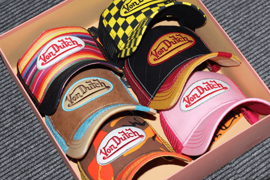 A collection of Von Dutch trucker hats in various bold colors and patterns, neatly arranged in a box. The hats feature the iconic embroidered Von Dutch logo, with designs including rainbow stripes, pink, camouflage, black with red stitching, and a yellow checkered pattern. The nostalgic Y2K fashion accessory makes a statement with its vibrant, retro aesthetic.